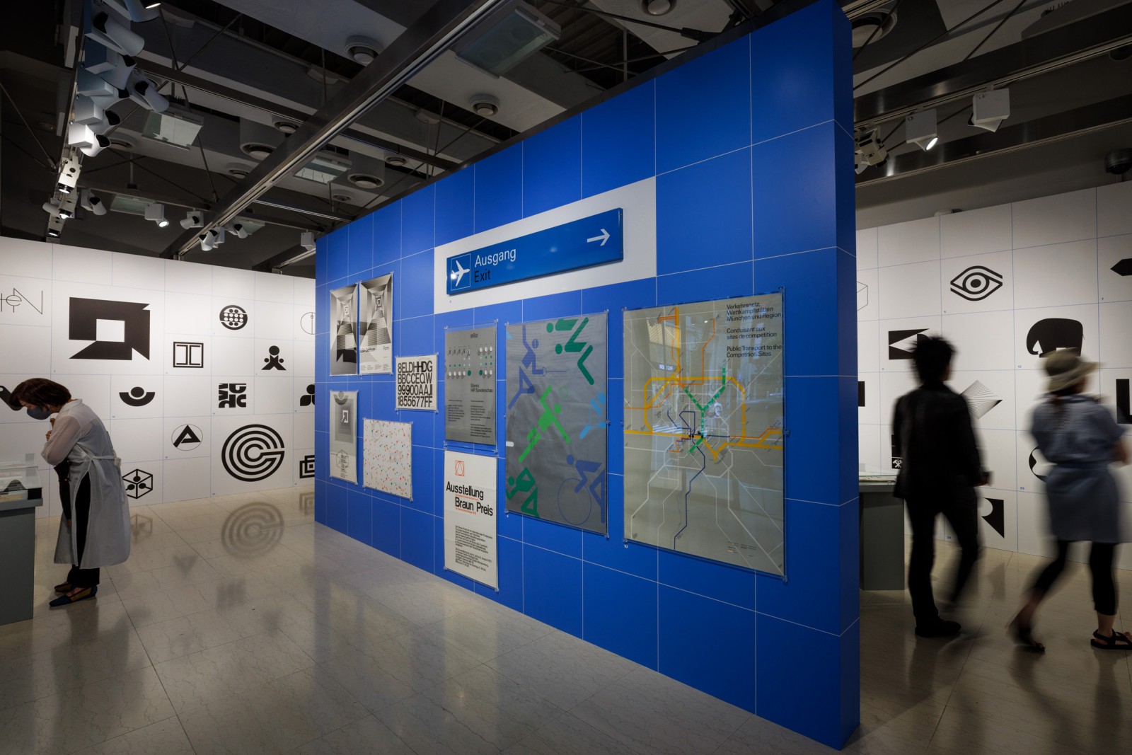 Ausstellungsansicht mit blauer Stellwand mit Highlight-Plakaten aus der A5 Collection, einem Flughafen Wegweiserschild, historischen Piktogrammen und Besuchern im Hintergrund