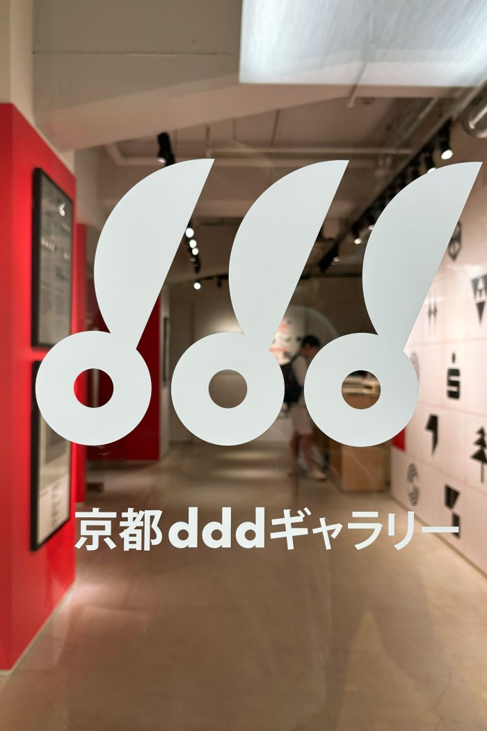 Eingangstür der Kyoto ddd Gallery mit weißem ddd-Logo und japanischer Beschriftung, dahinter die Ausstellung „Identity Systems Germany" mit roter Wand und Piktogrammtafel