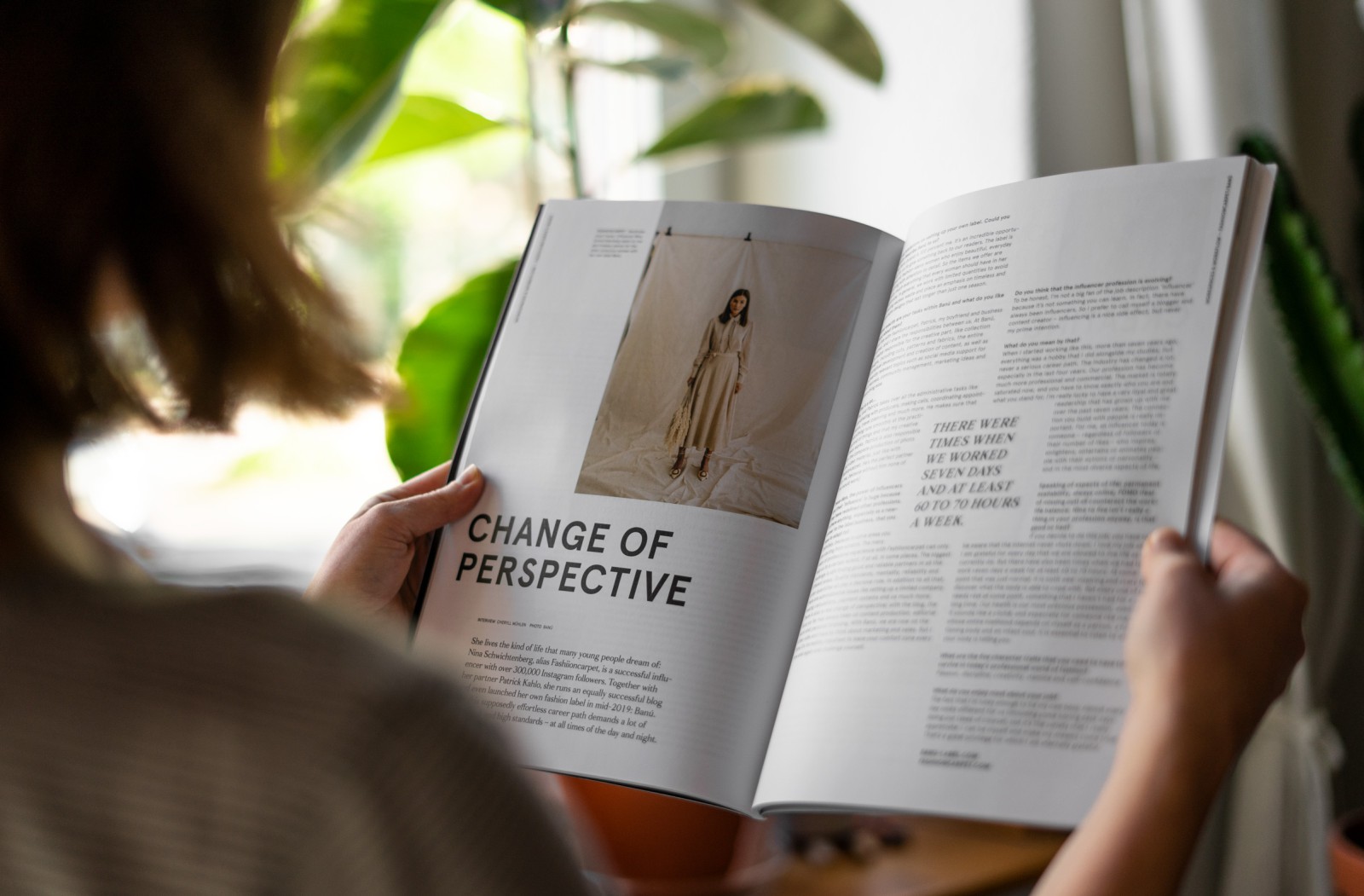 Atmosphärisches Foto einer Person, die ein aufgeschlagenes J'N'C Magazin vor einem Fenster mit Zimmerpflanzen liest; sichtbare Doppelseite zeigt das Titelthema „Change of Perspective" mit einem Modeporträt und mehrspaltigem Text