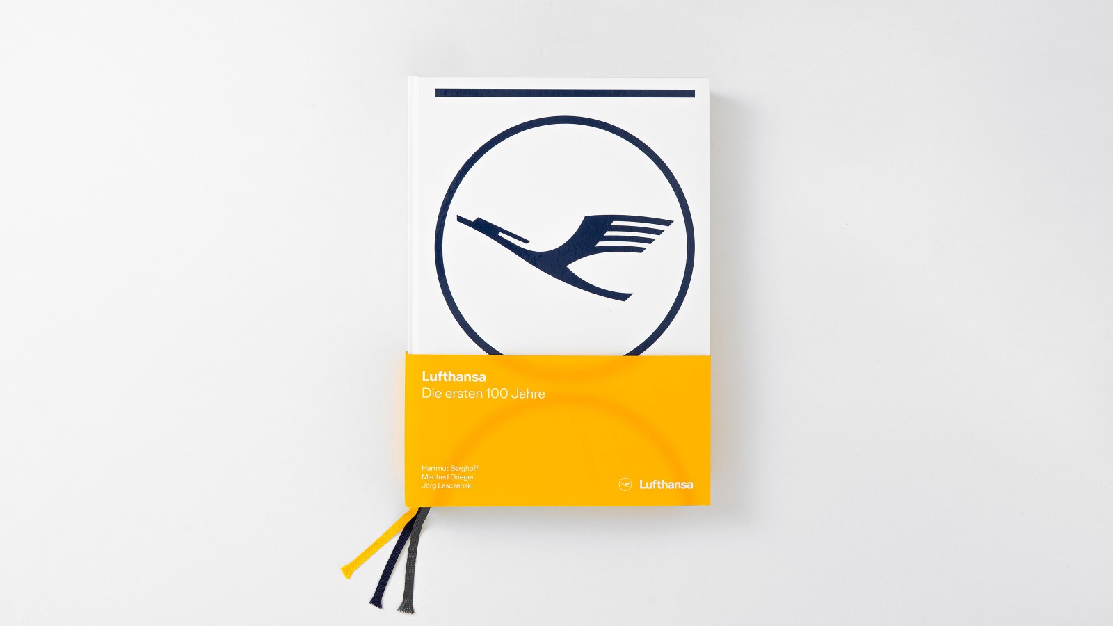 Vogelperspektive auf das Buchcover „Lufthansa – Die ersten 100 Jahre" (Prestel Verlag): weißer Einband mit dem klassischen Lufthansa-Kranich-Signet in Dunkelblau und einem gelben Streifen am unteren Rand mit Titel und Autorennamen Hartmut Berghoff, Manfred Grieger und Jörg Lesczenski