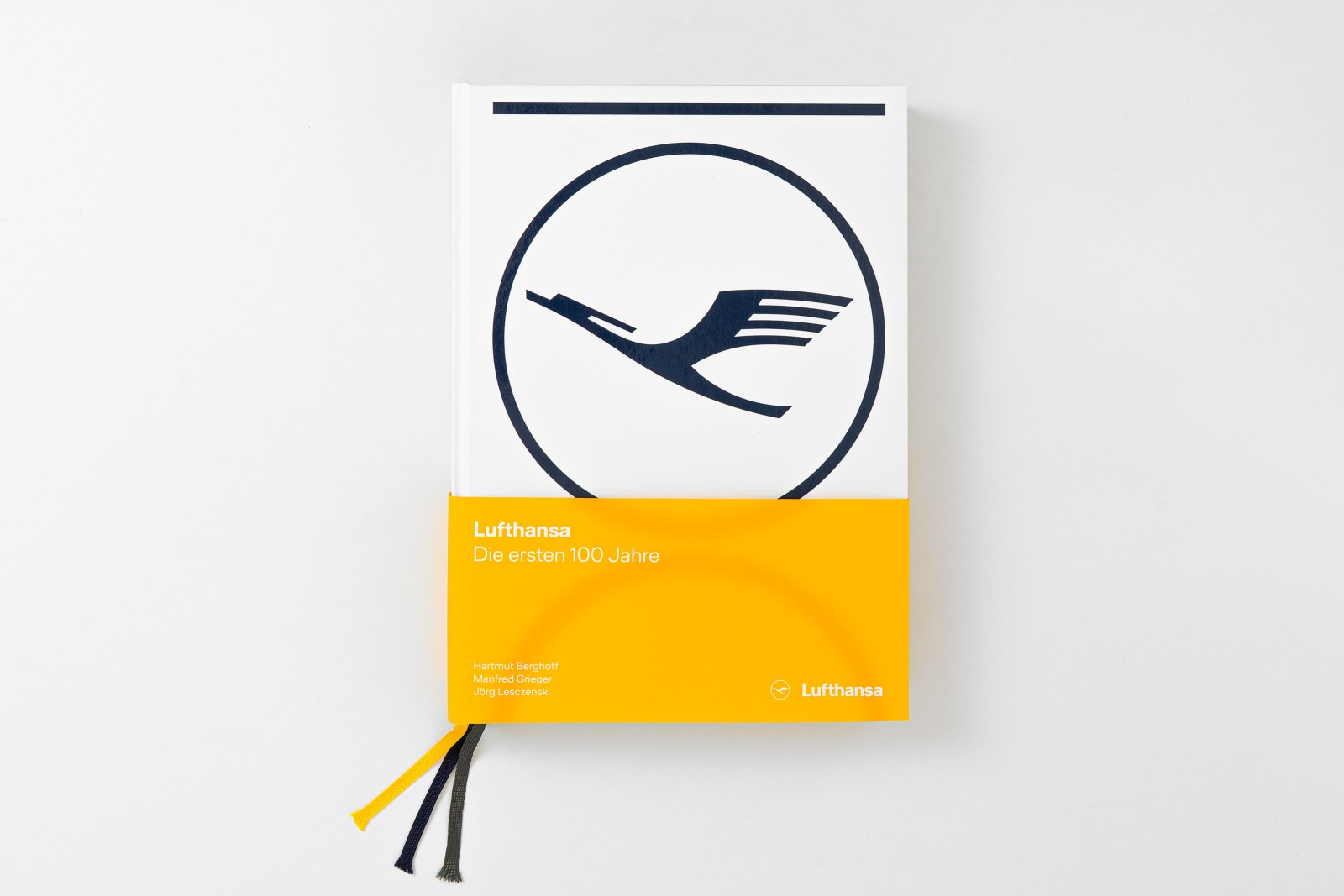Vogelperspektive auf das Buchcover &bdquo;Lufthansa &ndash; Die ersten 100 Jahre" (Prestel Verlag): wei&szlig;er Einband mit dem klassischen Lufthansa-Kranich-Signet in Dunkelblau und einer gelben Banderole