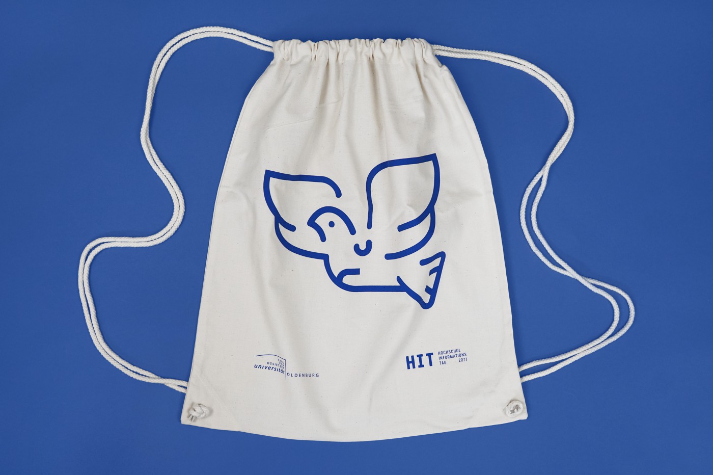 Heller Baumwoll-Turnbeutel mit blauer Linienzeichnung eines stilisierten fliegenden Vogels, Leuchtturms und Fahrrads sowie dem Logo der Carl von Ossietzky Universit&auml;t Oldenburg und dem HIT-Schriftzug 2017 auf blauem Hintergrund